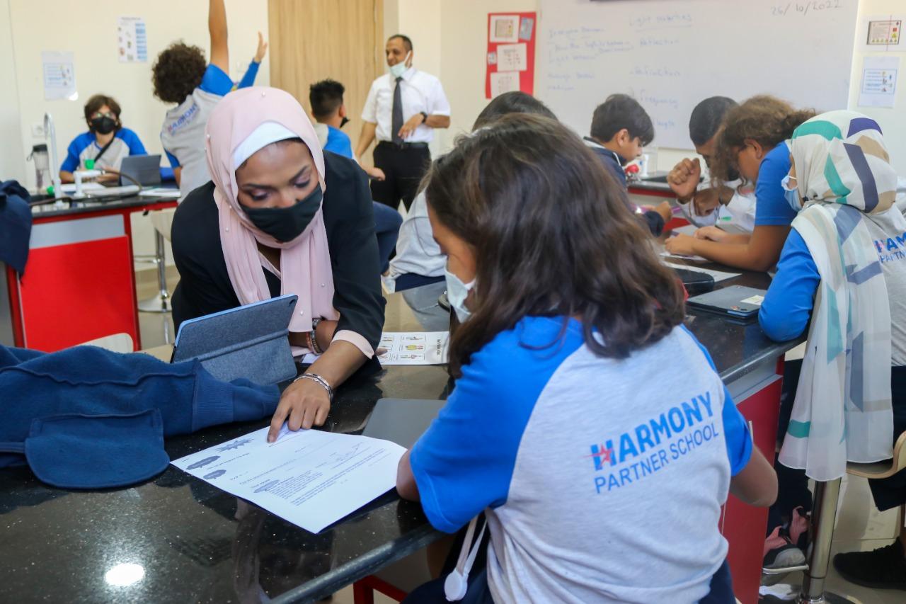 A classroom setting at IVY STEM International School, showing students and teachers engaged in educational activities. The image portrays a scene during a period of health precautions, with individuals wearing masks. A woman wearing a hijab and a face mask is seen interacting with another person, possibly assisting or discussing a document. The classroom environment includes a whiteboard, educational posters, and personal belongings on the tables.