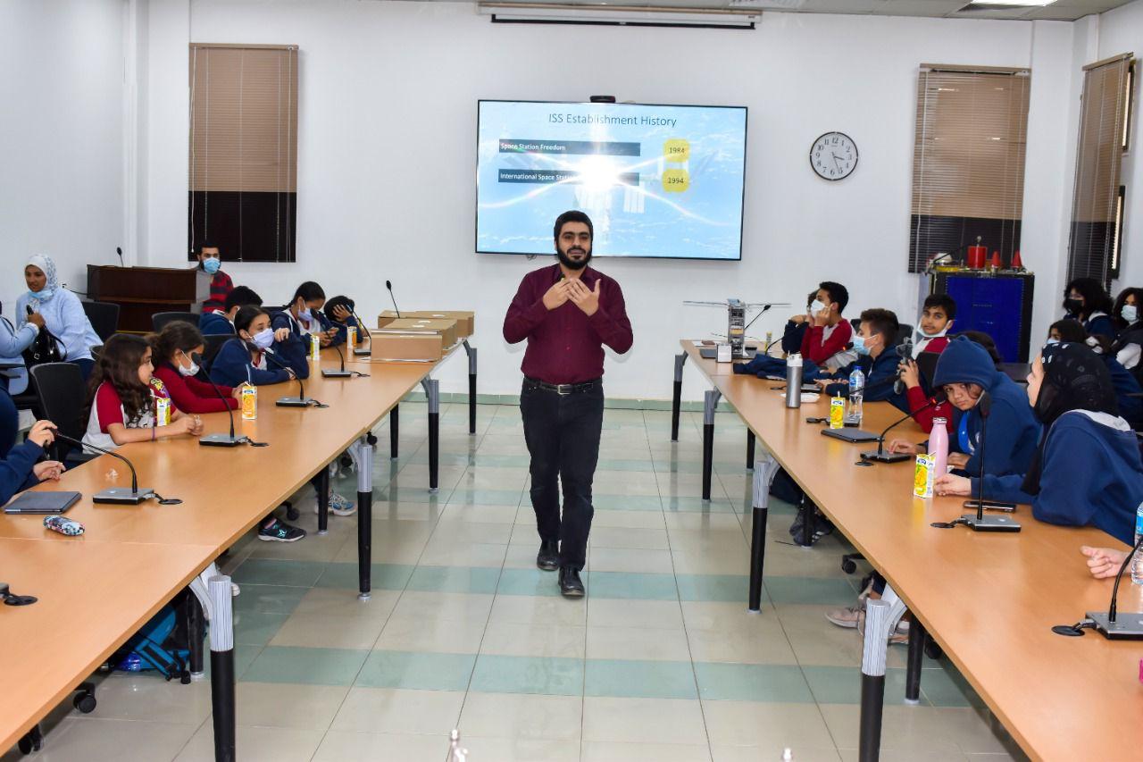 An indoor classroom or conference room at IVY STEM International School, where a lesson or presentation on space-related history is taking place. The instructor is addressing the audience, and a slide titled 'ISS Establishment History' is displayed on a large screen. Students are attentively listening and engaging in the session, some wearing face masks as a precautionary measure.