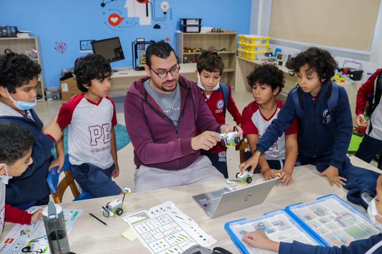 The image depicts a classroom at IVY STEM International School where a teacher conducts an interactive robotics lesson. Students in school uniforms gather around a table, observing a small robot held by the teacher. The table displays various teaching aids, including coding instructions and a laptop, emphasizing the use of technology. The room is decorated with STEM-related wall art, reflecting the school's focus on science, technology, engineering, and mathematics education.