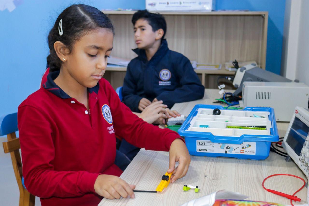 Two students engaged in hands-on STEM activities in an IVY STEM International School classroom. The girl is working with a STEM kit while the boy observes, both wearing school uniforms with the school emblem.