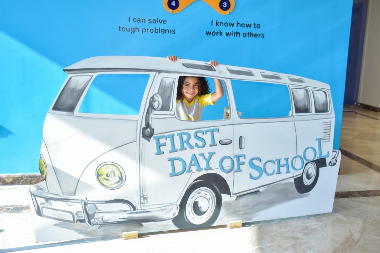 A life-size cutout with a painted illustration of a vintage-style bus with the text 'FIRST DAY OF SCHOOL' on the side. A young person is poking their head through a hole where the driver's window would be, giving the fun illusion that they are driving the bus. Positive affirmations 'I can solve tough problems' and 'I know how to work with others' are displayed in textual bubbles. This setup represents a first day of school photo opportunity at IVY STEM International School.