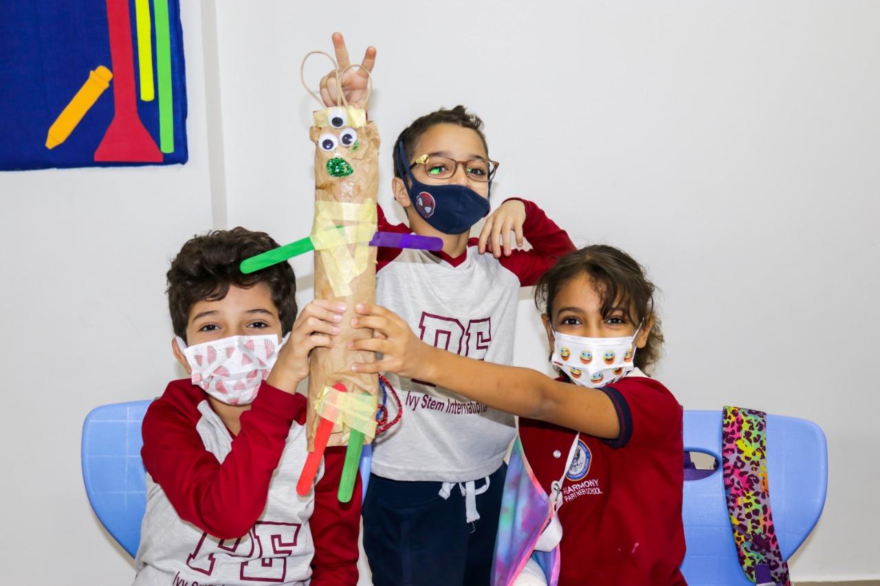 Children at IVY STEM International School wearing face masks and proudly displaying their homemade art project made from a cardboard tube. The project is adorned with colored papers, googly eyes, and pipe cleaners, reflecting the students' creativity and learning in a classroom setting.