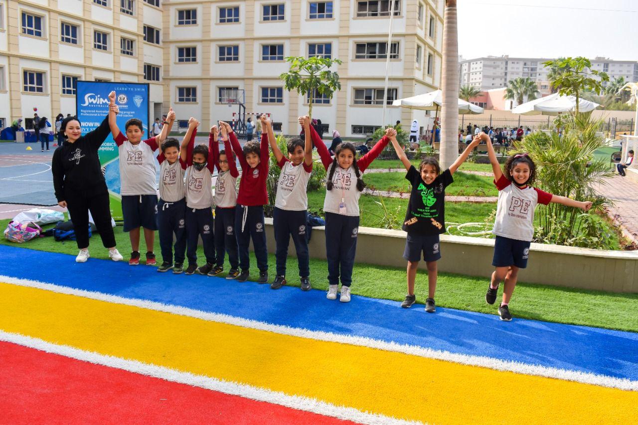 Group of children participating in a lively and cheerful physical education class or school event at IVY STEM International School. They are lined up outdoors on a colorful, striped floor surface, with their arms raised up forming a chain. The children are wearing school or athletic attire, and a woman, likely an instructor or teacher, is seen participating with them. The background includes a school-like building, grassy areas with small trees and plants, and a partially visible basketball hoop, indicating the area is equipped for recreational activities.