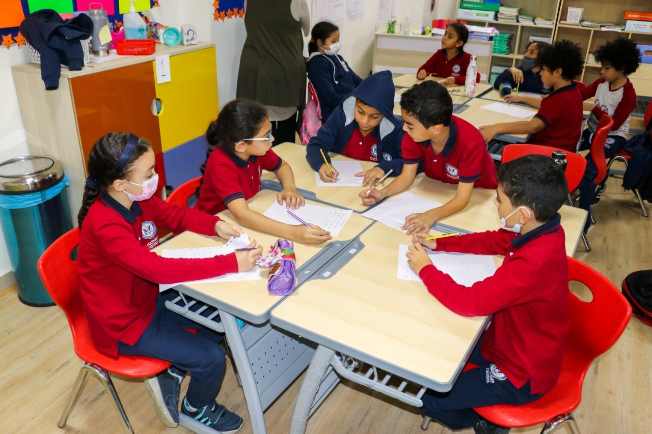 A vibrant classroom setting at IVY STEM International School, with students engaged in educational activities. The children are dressed in school uniforms, wearing protective face masks. The atmosphere is lively, with colorful decorations and interactive learning. A teacher is present, observing or assisting the students.