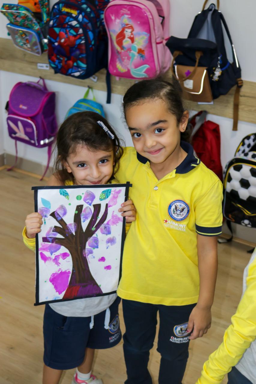 Two young students from IVY STEM International School engaged in an art activity in a classroom. They are holding a handmade artwork featuring a colorful tree with purple and pink leaves. The children are smiling and proud, showcasing their accomplishment and enjoyment in the art-making experience.