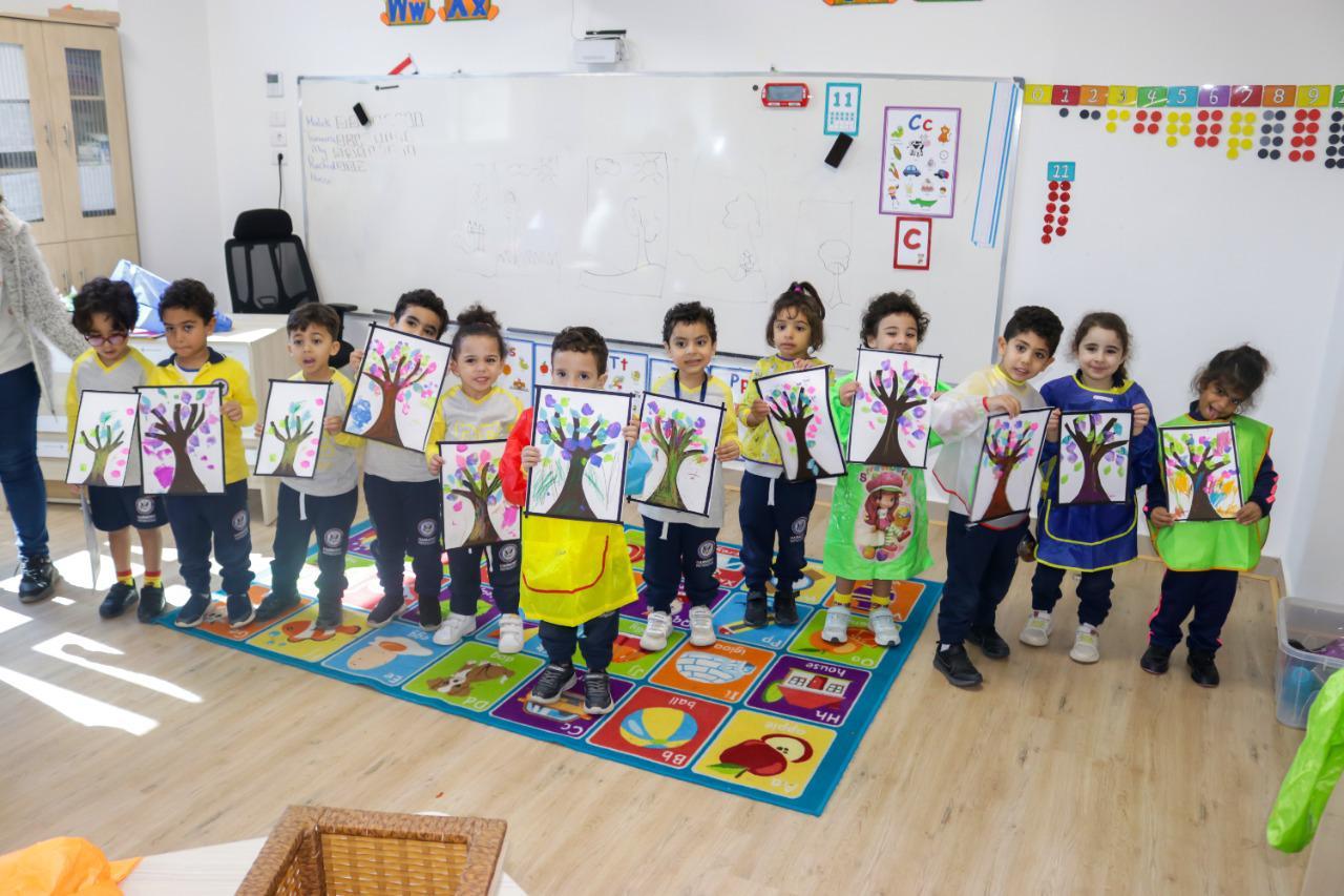 This image captures young children at IVY STEM International School showcasing their creativity through colorful handprint tree paintings. The classroom environment is filled with educational materials, including a whiteboard, posters, and a vibrant alphabet carpet, reflecting the school's commitment to fostering artistic expression and learning.