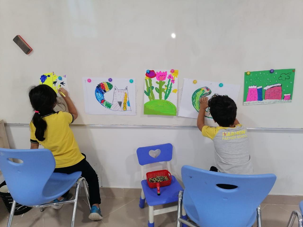 Children engaged in a creative activity in a classroom setup at IVY STEM International School. They are working on colorful drawings displayed on the white wall, showcasing artistic expression and educational engagement.