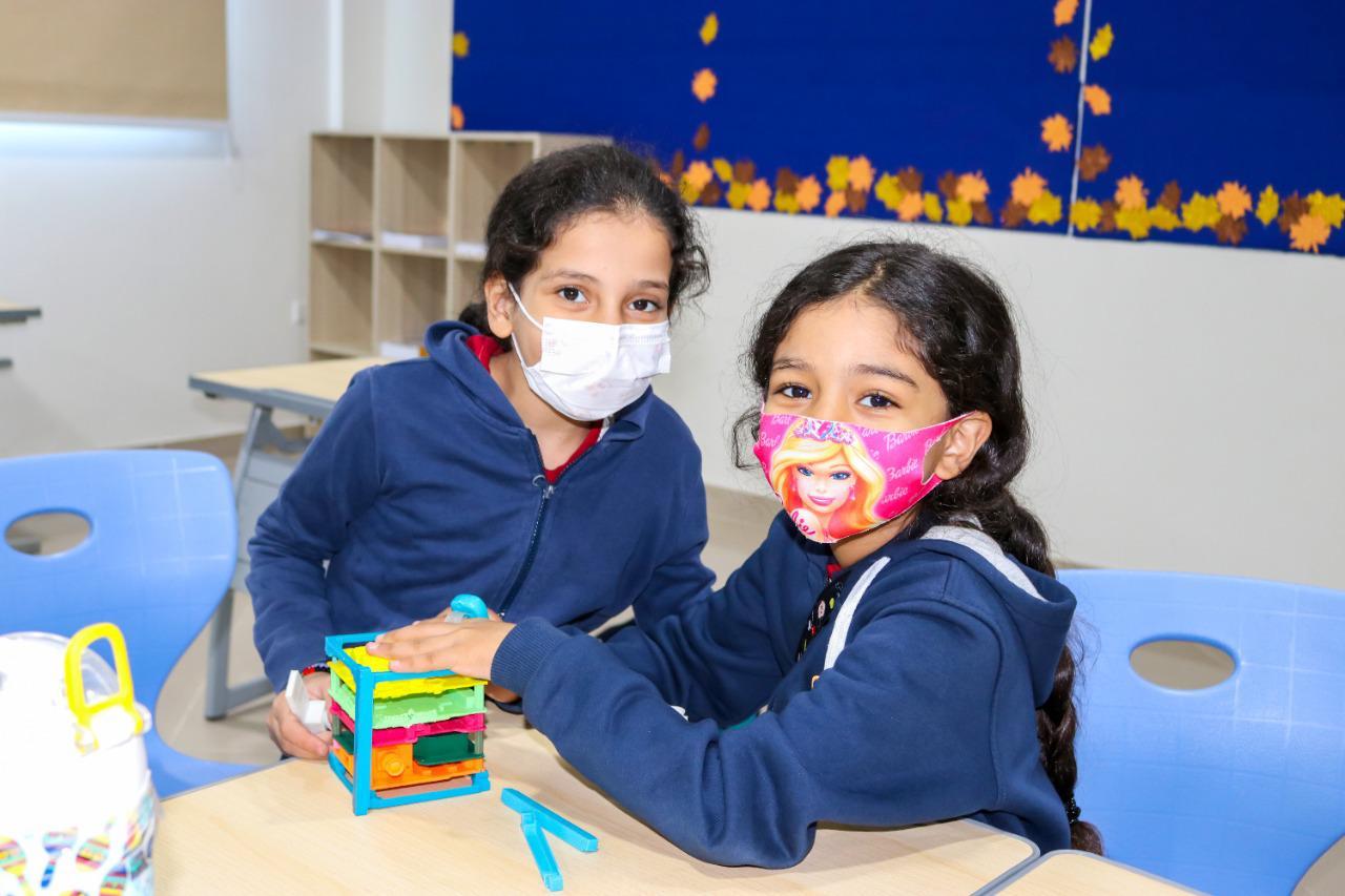 Two young girls wearing school uniforms and face masks are seen in a classroom at IVY STEM International School. They are engaged in educational activities with a colorful toy structure on the table. The classroom is well-organized with fall-themed decorations on the wall.