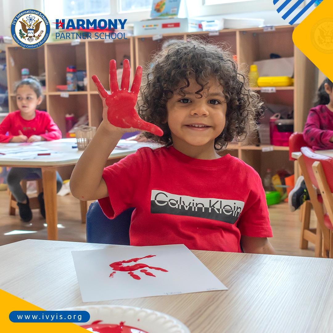 A young child at IVY STEM International School engaged in a painting activity, showcasing creativity and joy in the classroom. The child proudly displays a red handprint artwork, symbolizing the school's commitment to fostering artistic expression and educational development. This image represents the vibrant and internationally minded environment of IVY STEM International School, a Harmony Partner School affiliated with the International Baccalaureate (IB) program.