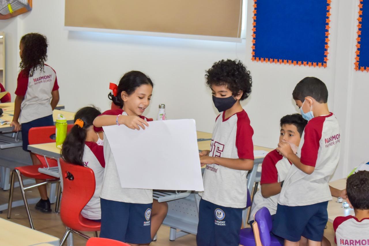 A vibrant classroom scene at IVY STEM International School with students engaged in interactive learning activities, including a girl holding up a white paper and discussing it with a boy. The classroom is well-lit with natural light, modern furniture, and colorful decorations on the walls.
