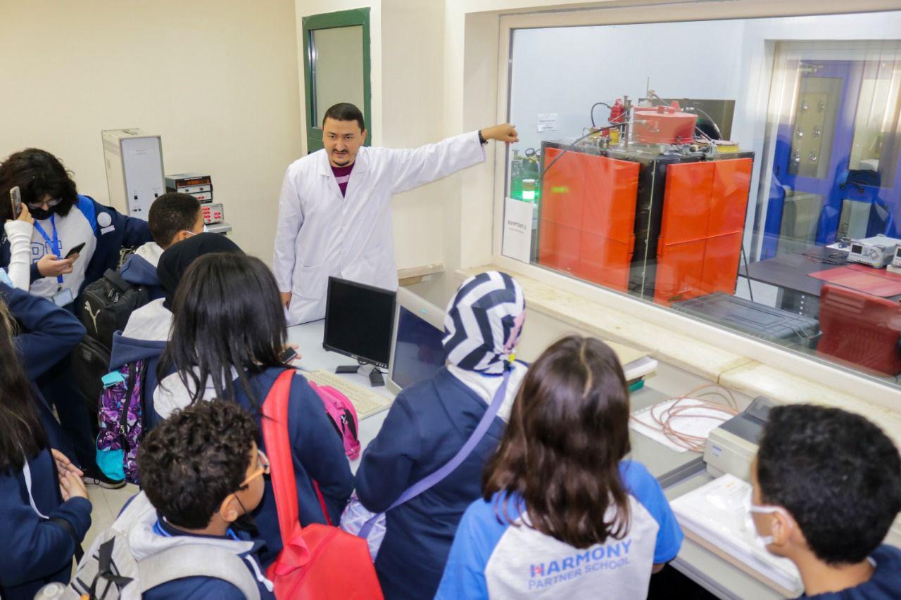 In this image, students at IVY STEM International School are engaged in a classroom science experiment. A teacher or scientist in a white lab coat is demonstrating the use of technical equipment, while students of various ages observe and participate. The scene showcases interactive learning and real-world applications of scientific principles.