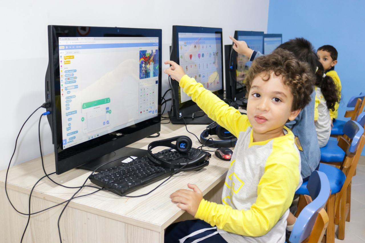A young child in a classroom at IVY STEM International School engaged in a computer class, surrounded by other students. The child is wearing a yellow long-sleeved shirt with a denim vest and is seated at a computer workstation, pointing at the screen. The environment reflects the school's modern approach to education, emphasizing technology and computer literacy.