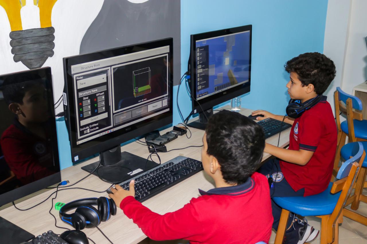 The image showcases students at IVY STEM International School engaged in a digital project or game in a computer lab. They are using 3D modeling software and block-style graphics, demonstrating their creativity and problem-solving skills. The educational environment is enhanced by the presence of headphones and educational posters on the walls.