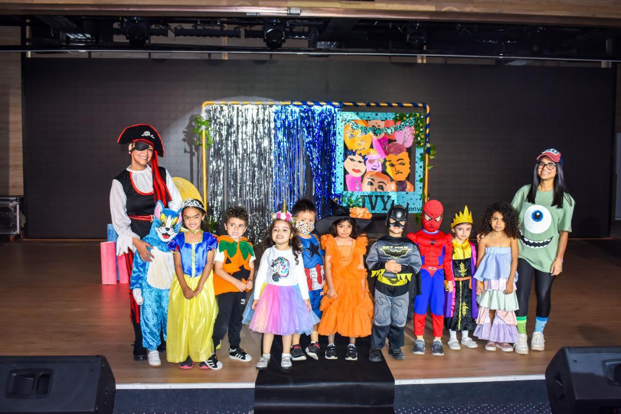Group of children and adults dressed in various costumes at a festive event at IVY STEM International School. The costumes include popular characters from fiction, such as superheroes and classic fairy tale figures. The backdrop features the name 'IVY' on a colorful banner.
