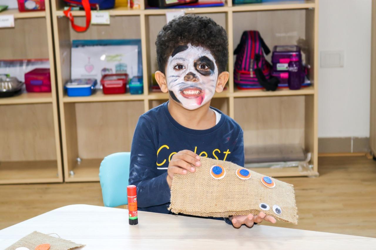 A child at IVY STEM International School engaged in a craft activity, holding a piece of burlap fabric with googly eyes attached. The child's face is painted in black and white, resembling a panda or similar animal face painting design.