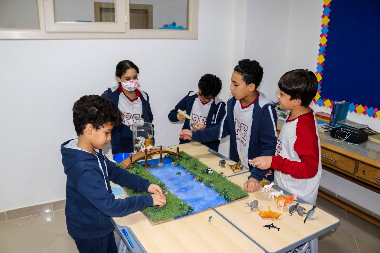 Group of schoolchildren engaged in an educational activity at IVY STEM International School. They are learning about ecosystems and habitats through a diorama. The children are wearing school uniforms with the logo and acronym 'PPE' visible, indicating hands-on learning and teamwork. The photo was taken during a time of heightened health precautions, possibly during the COVID-19 pandemic.