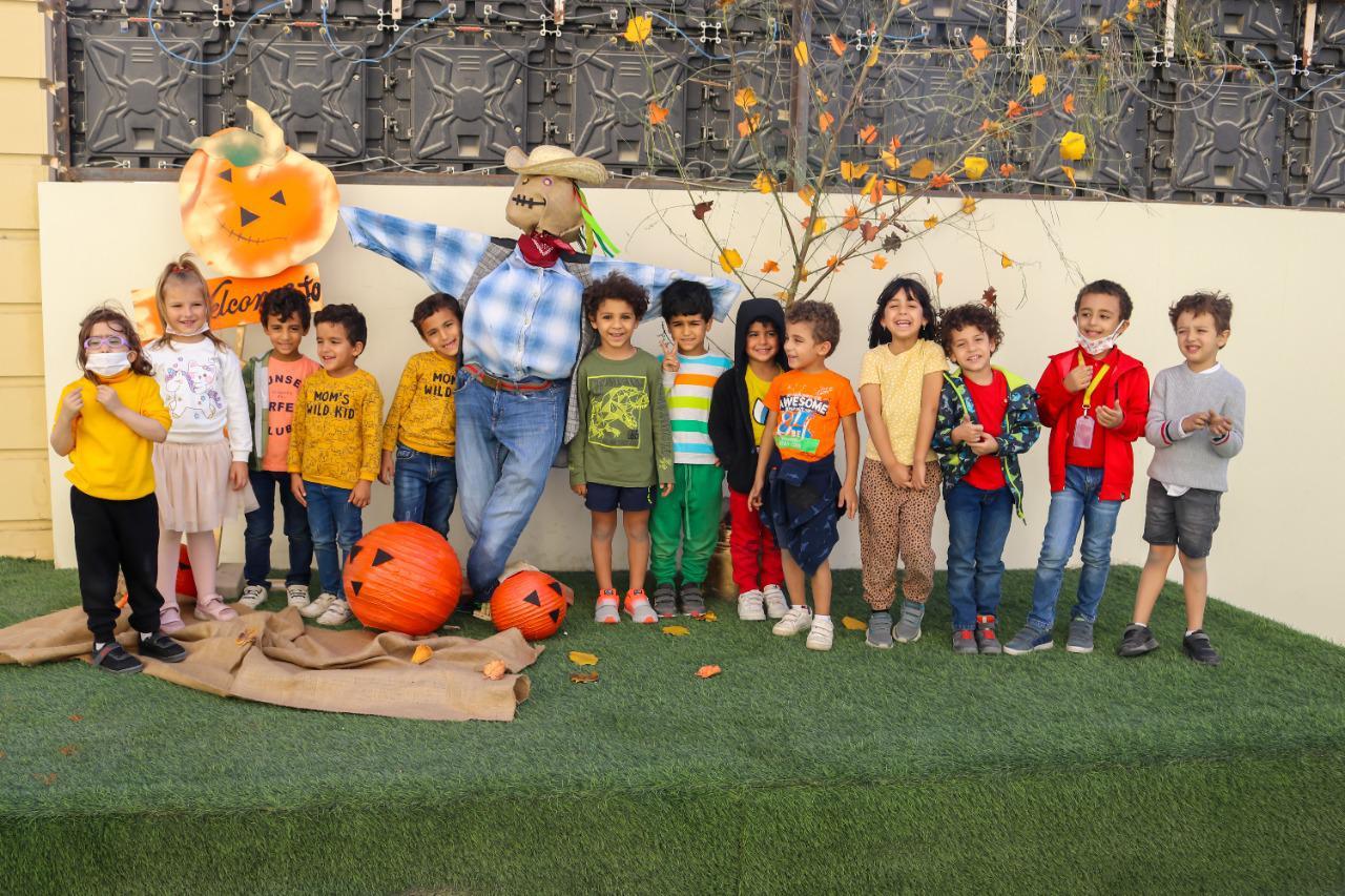 A group of children from IVY STEM International School posing for a photo during a Halloween themed event. The children are standing on a grassy surface with Halloween decorations, including a scarecrow figure, pumpkin decorations, and a tree with autumn leaves. They are wearing festive attire, creating a cheerful and celebratory atmosphere.