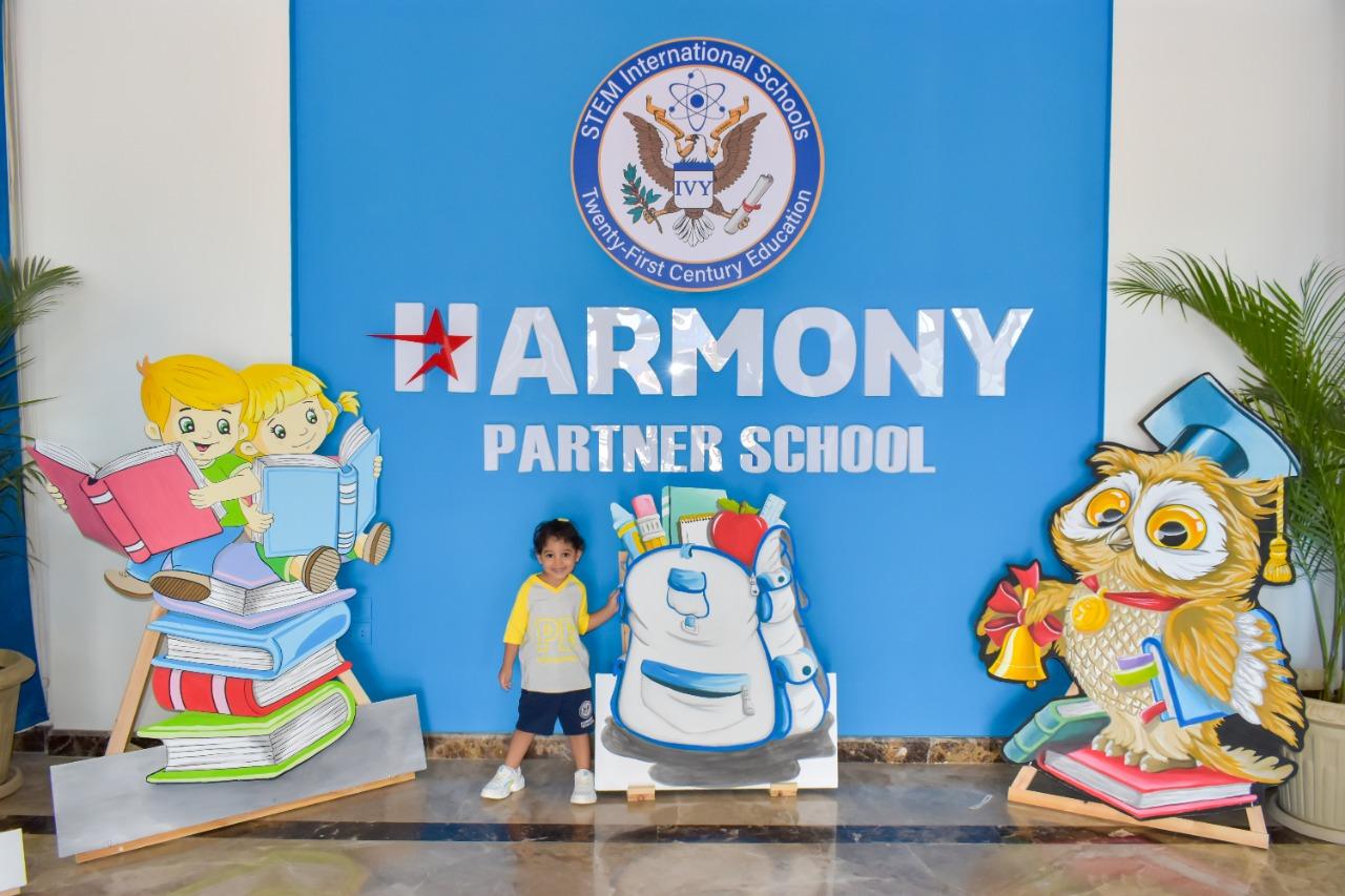 A young child standing in front of a colorful backdrop with the words 'HARMONY PARTNER SCHOOL' displayed prominently. Educational theme with animated children characters sitting on books and reading, and a cartoonish owl wearing a graduation cap and holding a diploma and a bell. Blue wall with a logo reading 'STEAM International Schools' and 'Twenty First Century Education' with an eagle emblem. Represents IVY STEM International School's partnership with Harmony Partner School and commitment to providing a modern educational environment.