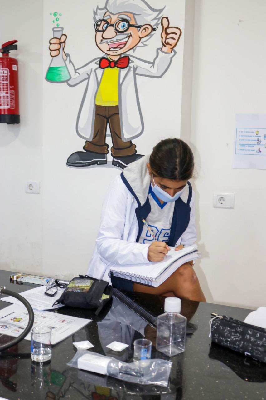 An image showcasing a student engaged in laboratory work at IVY STEM International School. The student is wearing a school uniform and lab coat, demonstrating the school's focus on practical science education. The laboratory setting includes various materials, worksheets, and a playful illustration of a mad scientist, highlighting the school's fun and educational approach to science.