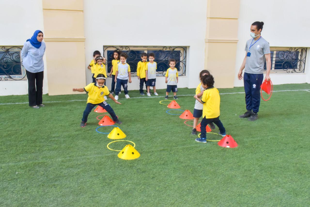 Children from IVY STEM International School participating in an outdoor activity, wearing matching yellow tops. They are supervised by adults and navigating through an obstacle course or play area with orange cones and yellow hoops. This activity promotes physical activity and coordination among the students.