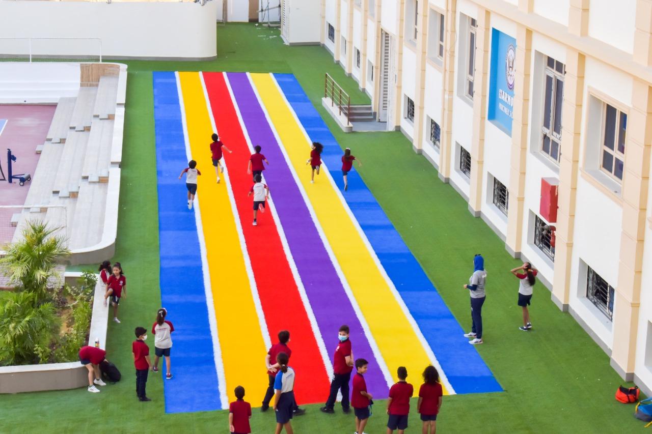 This image showcases the vibrant outdoor area of IVY STEM International School. It features a rainbow-colored track where children in school uniforms engage in various activities, including running and playing. The symmetric architecture of the surrounding building with multiple windows and a minimalistic design adds to the aesthetic appeal. Benches are available for students to sit and relax. The elevated perspective of the image provides a comprehensive view of the entire area.