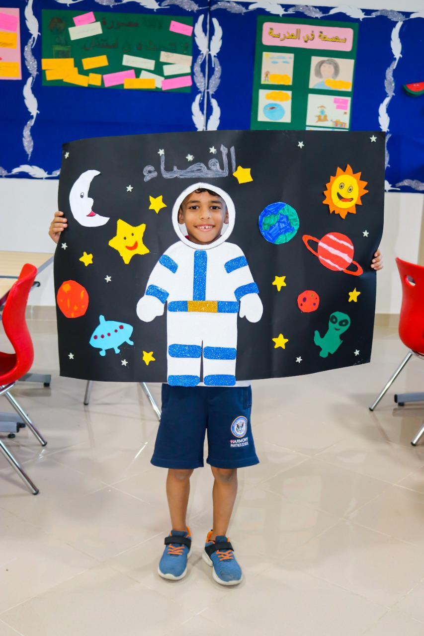Young child participating in a space-themed activity at IVY STEM International School, dressed as an astronaut and holding a poster with celestial elements. Educational posters and bright red chairs can be seen in the background.