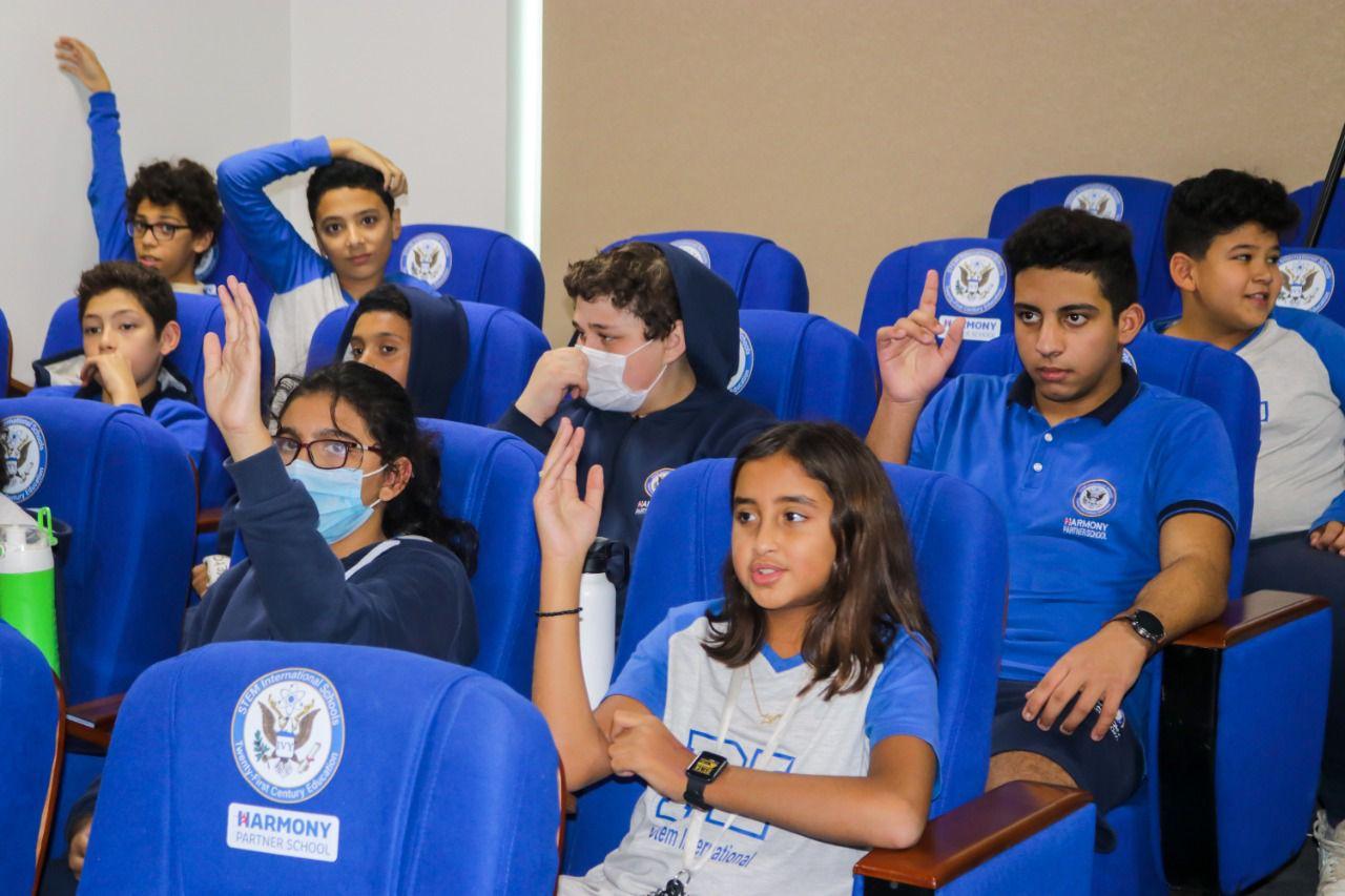 Group of students from IVY STEM International School participating in an interactive classroom event. Students are seated in rows of blue chairs with the emblem of the Harmony Partner School. Various ages and attire suggest a diverse student body engaged in educational activities.