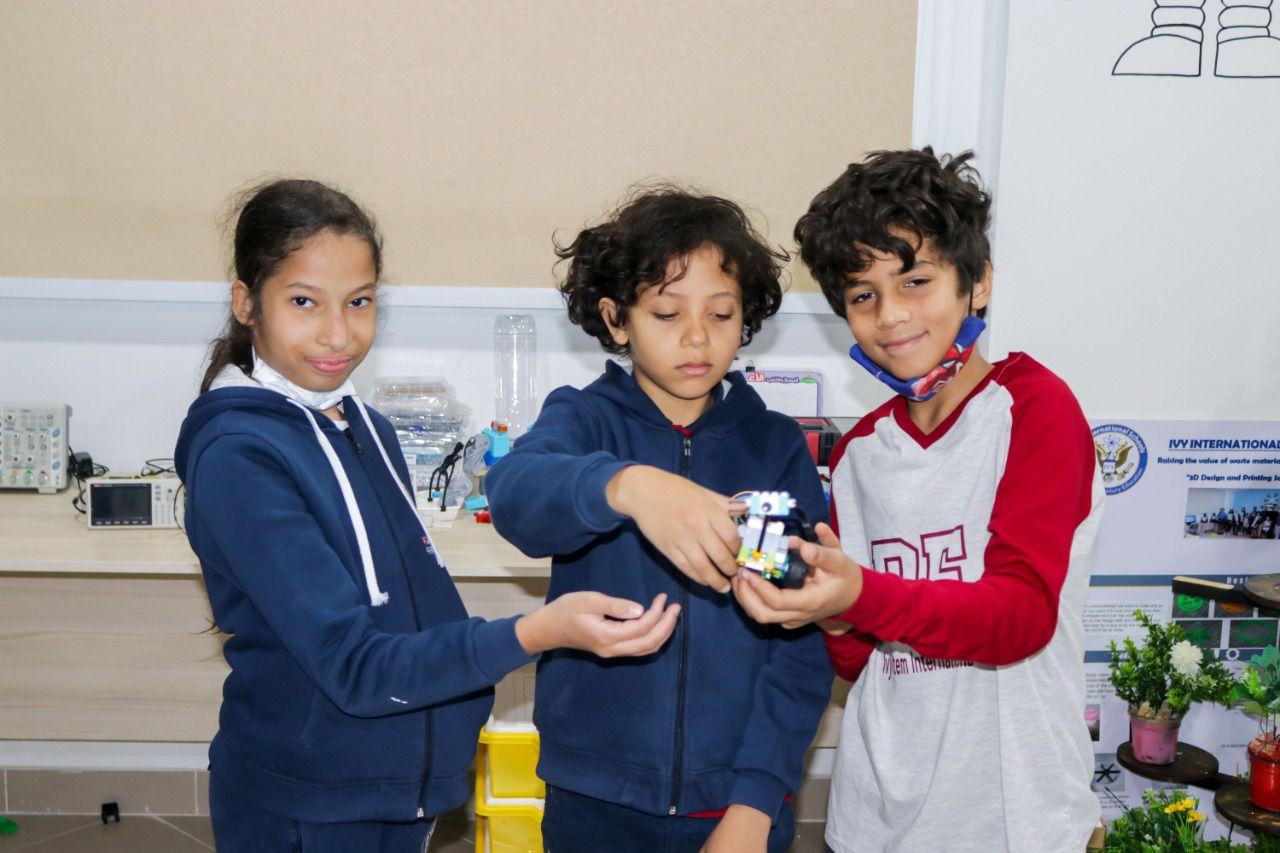 Three young students from IVY STEM International School engaged in a robotics learning activity in a classroom or lab setting. The students are seen using educational construction kits and a small robot, demonstrating the school's commitment to hands-on STEM education.
