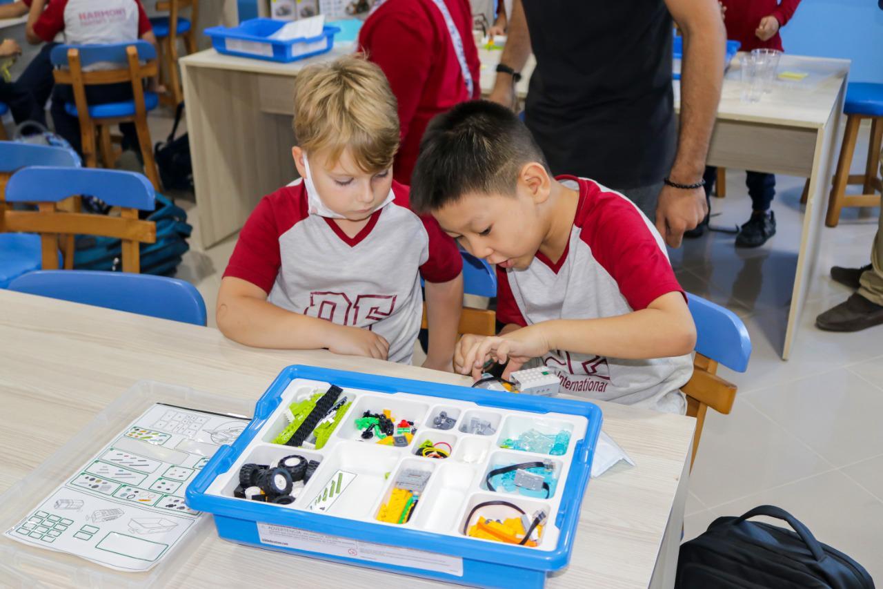 Two young students from IVY STEM International School are seen working together on a robotics kit. They are engaged in assembling a model or a simple machine using various components like wheels, gears, and colorful parts. This educational activity takes place in a well-lit classroom environment, indicating the school's focus on collaborative learning.