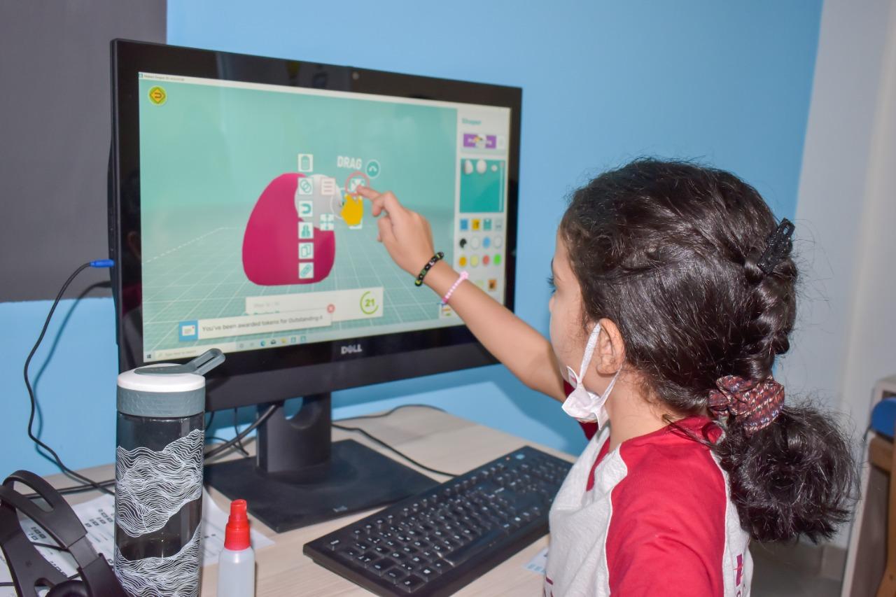 This image depicts a young girl actively participating in an educational program at IVY STEM International School. She is engrossed in an interactive activity involving shapes and colors on a computer monitor. The use of a face mask, along with a water bottle and hand sanitizer on the desk, reflects the school's commitment to health and safety measures during the COVID-19 pandemic. The image captures the engaging classroom or home study environment at IVY STEM International School.