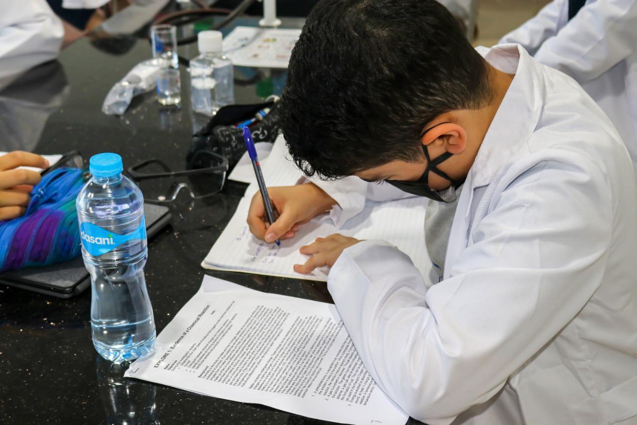 A student from IVY STEM International School conducting a scientific experiment in the laboratory, wearing a white lab coat and focusing on writing in a notebook.