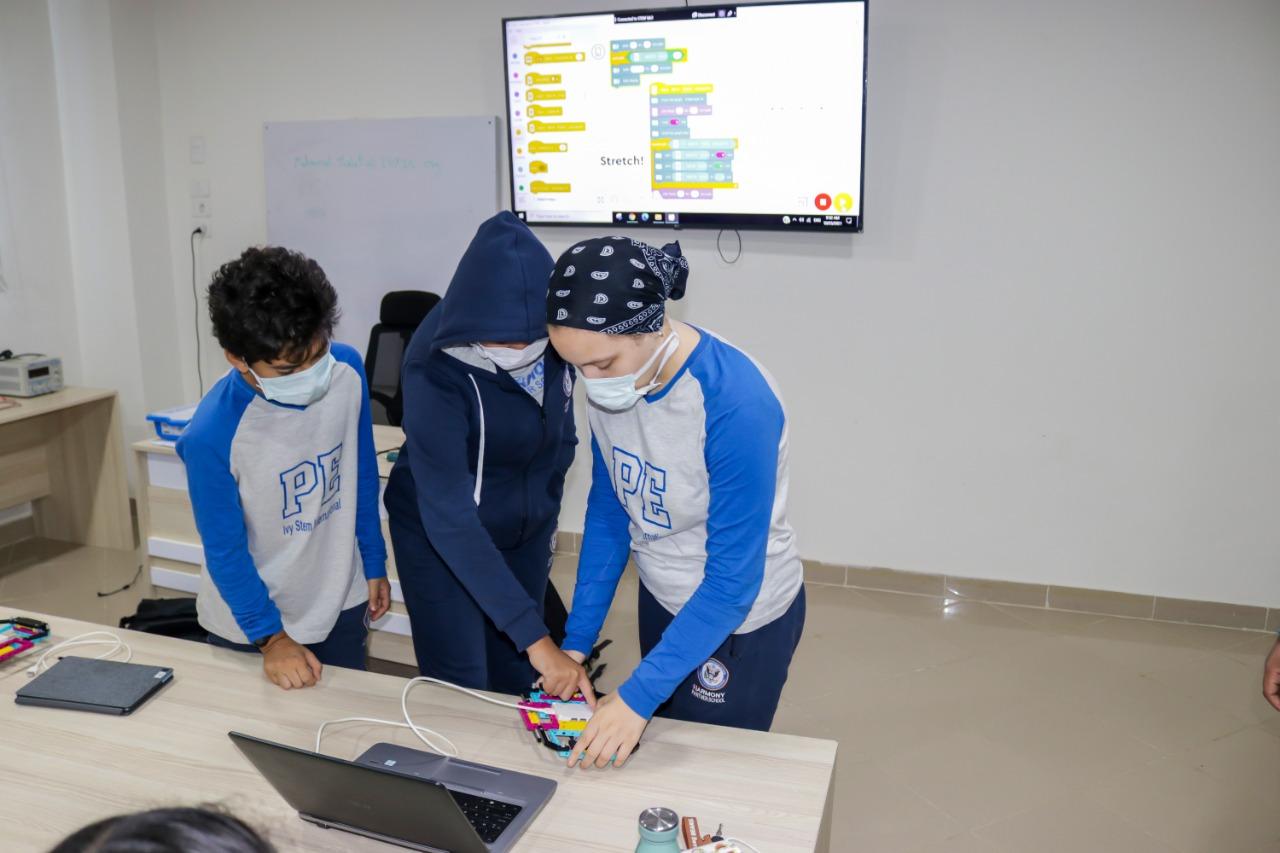 The image depicts students at IVY STEM International School participating in a robotics or STEM educational activity. They are engaged with a block-based robotic device connected to a laptop, learning programming and engineering concepts. The classroom setting reflects the school's commitment to modern and well-equipped learning environments.