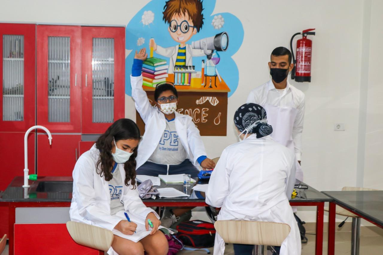 Students in a science classroom at IVY STEM International School wearing lab coats and masks, actively participating in hands-on learning activities. The classroom is equipped with science lab benches, educational materials, and safety measures.