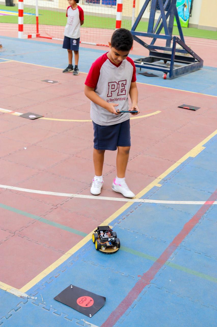 A young student at IVY STEM International School engaged in a robotics activity, controlling a small robot with a smartphone. The student is outdoors on a multi-colored court, wearing a PE uniform. Another student is also seen participating in a similar activity in the background.