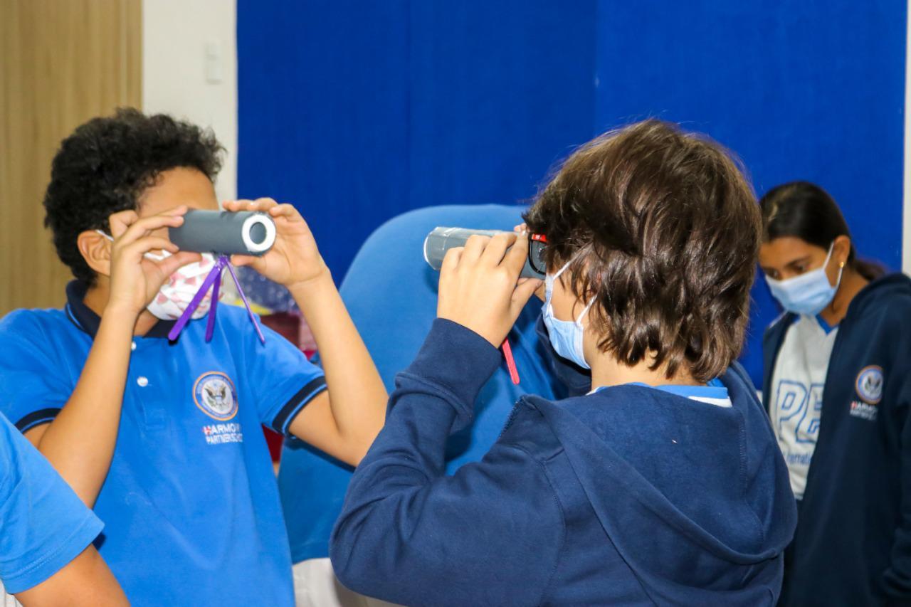 Two students from IVY STEM International School wearing masks and blue uniforms actively engaging in an educational activity with handmade kaleidoscopes or telescopes. A teacher or supervisor observes their progress in a classroom setting.