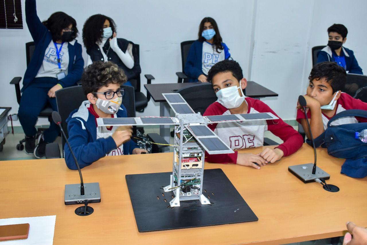 Students at IVY STEM International School actively engage in a classroom project involving a model satellite with solar panels. They are wearing face masks as a health precaution. The satellite model, constructed from metal frames, is the focus of this STEM lesson. The presence of a microphone suggests a presentation or facilitated discussion.